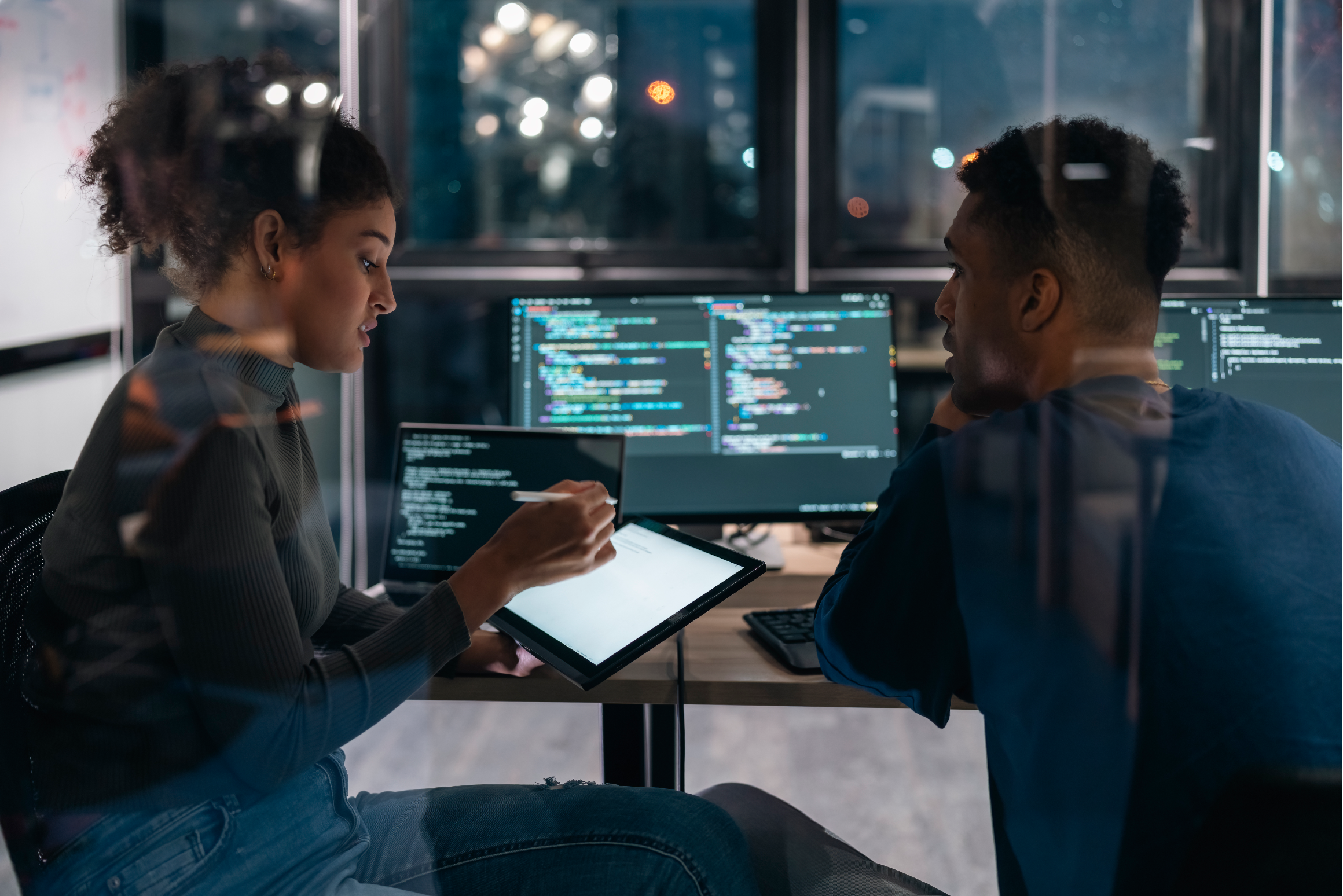 A software developer points to code on a tablet while a colleague looks on, with code displayed on monitors behind them.